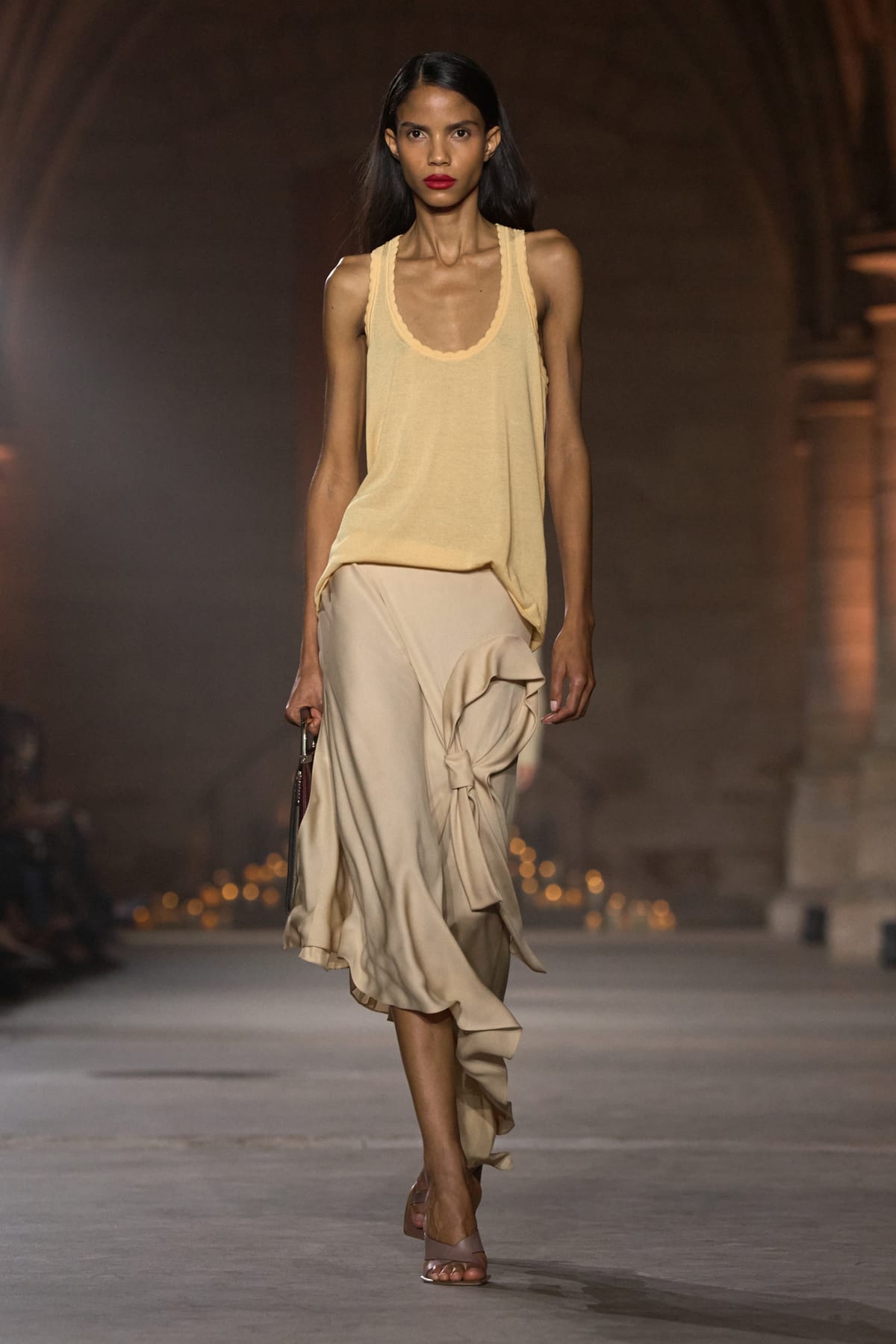 A Victoria Beckham Spring 2027 look: a pale yellow scoop-neck tank over a cream satin skirt gathered into a knot at the hip, with nude sandals